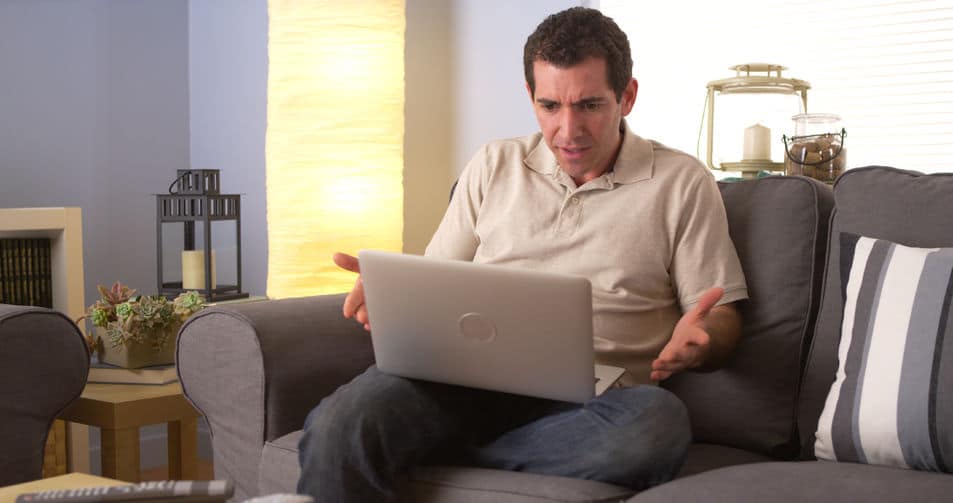 a frowning man sitting on the couch looking at his laptop