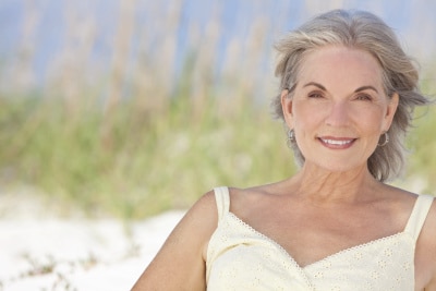 An attractive elegant senior woman sitting on a white sand beach with grass and a blue sky behind her.