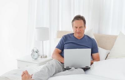 older man wearing blue shirt with laptop in bed