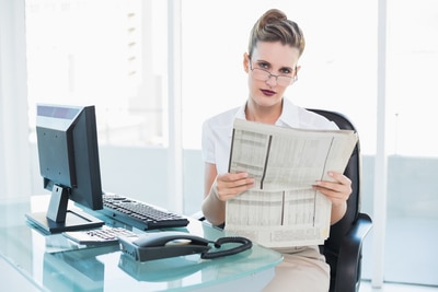 Serious businesswoman wearing glasses holding a newspaper in bright office