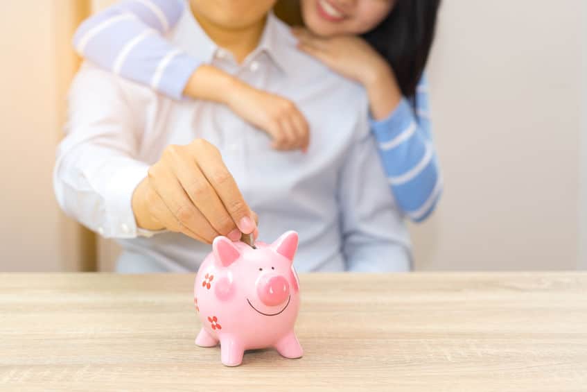 Smile couple putting a coin into a pink piggy bank on wooden desk - save money for the future