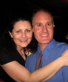 middle-aged man wearing blue longsleeves and tie, hugged by his wife