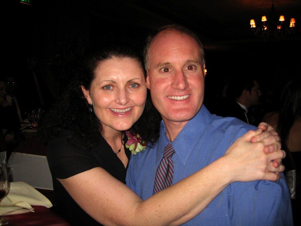 middle-aged man wearing blue longsleeves and tie, hugged by his wife